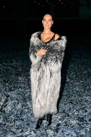 Model wearing a dramatic black and white shaggy faux fur coat stands on dark textured ground at night.