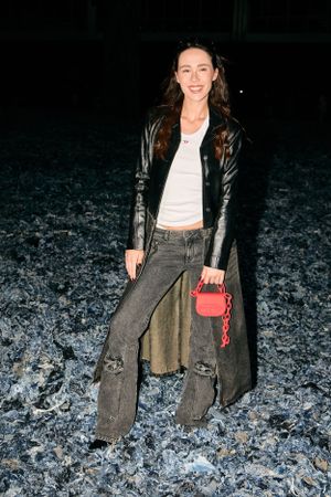 Casual street style outfit featuring leather jacket, ripped jeans and small red handbag on dark background.
