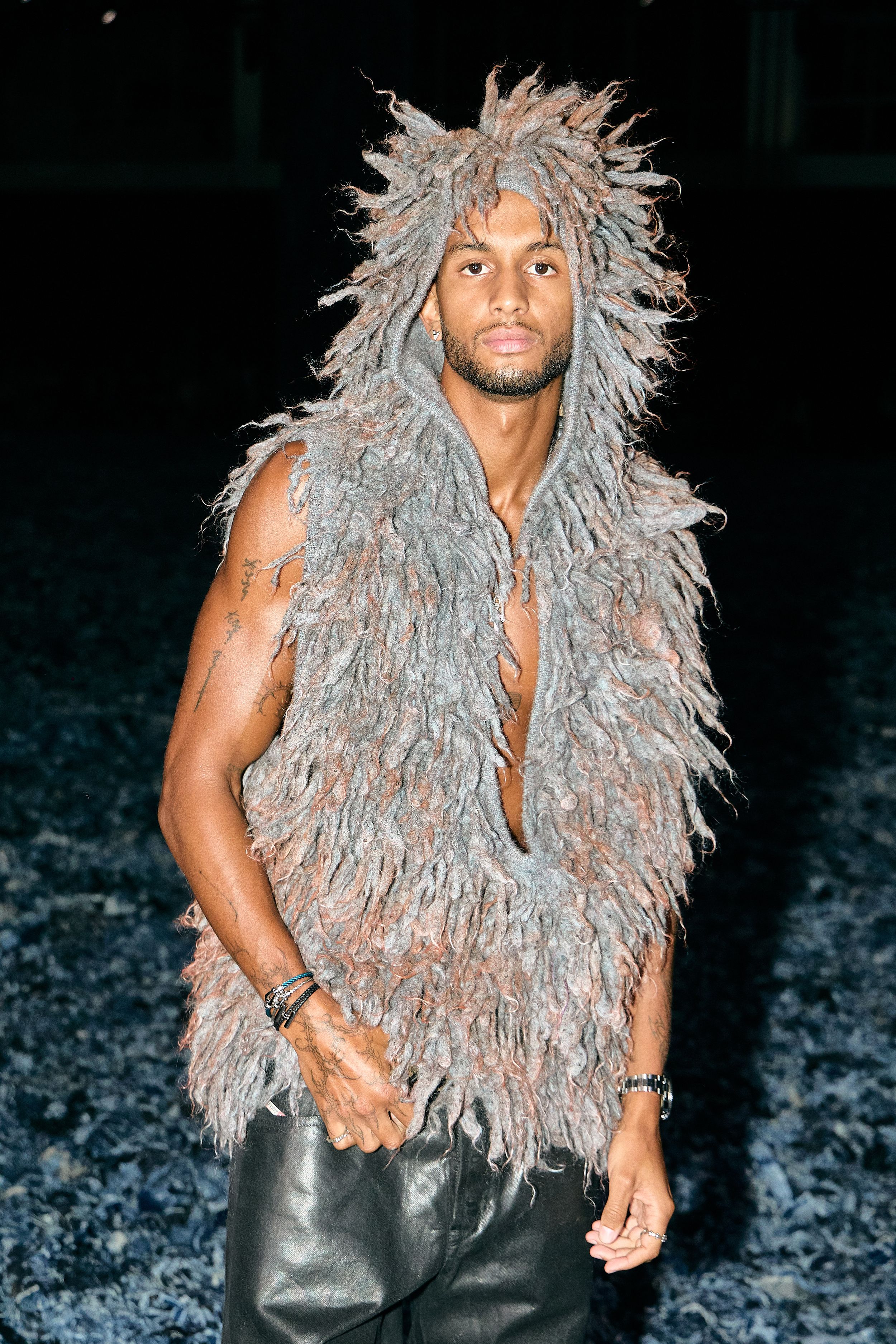 Person wearing shaggy grey faux fur vest with hood and leather pants against dark background.