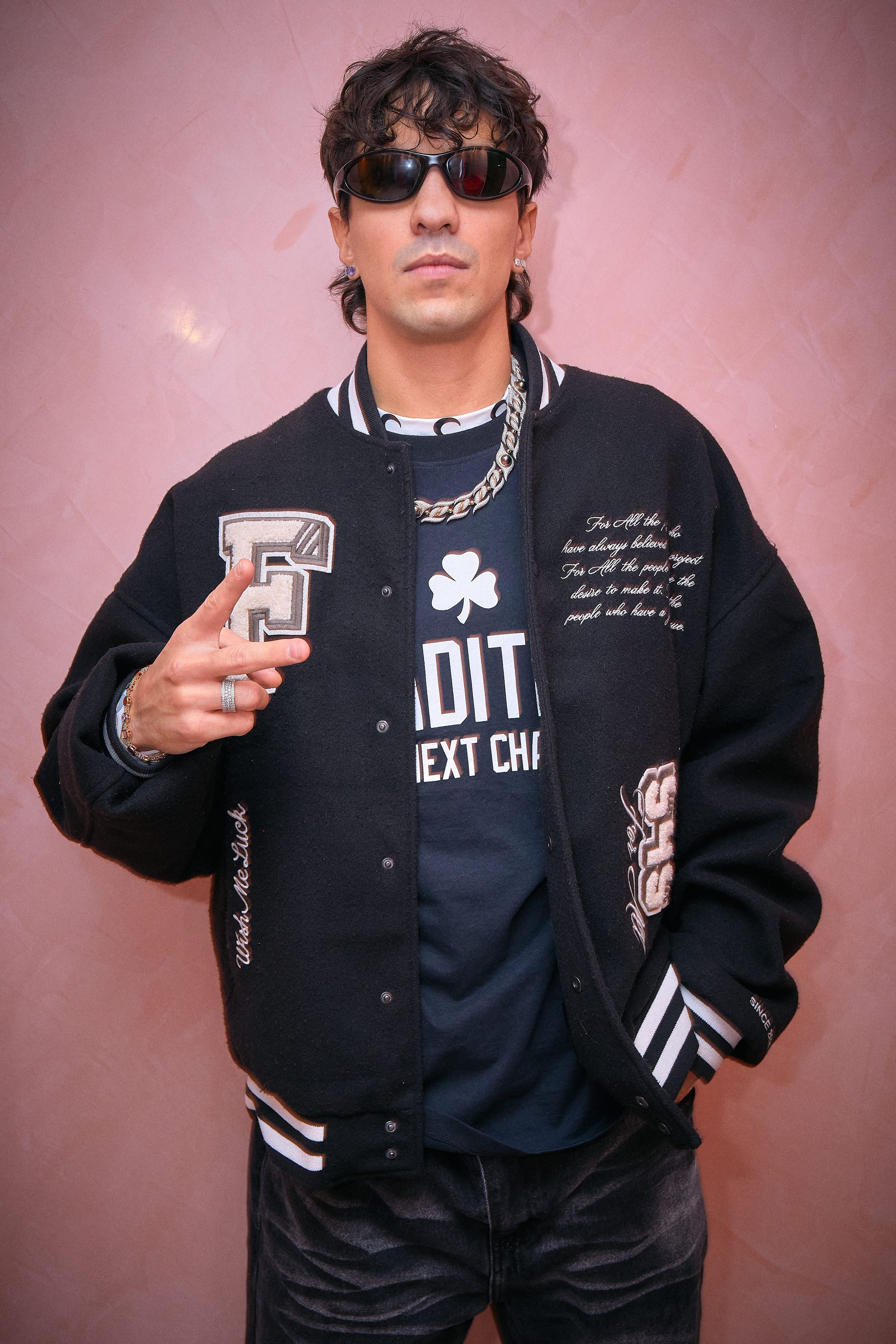 Streetwear model poses against pink wall wearing varsity jacket and silver chain necklace.