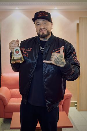Performer in black jacket holds up bottle and gestures while standing in front of orange couch.