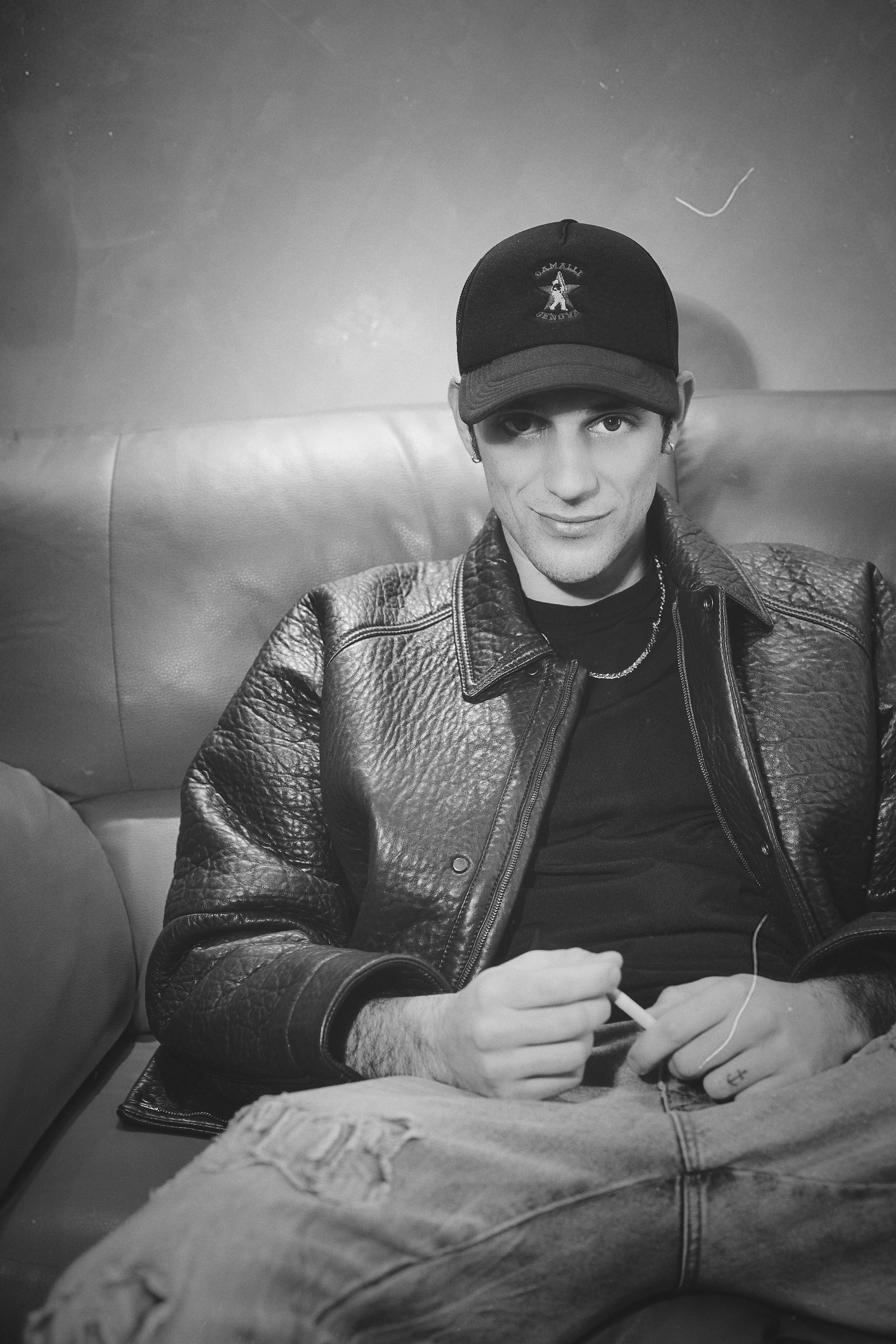 Monochrome portrait of person in leather jacket and baseball cap sitting casually on couch.