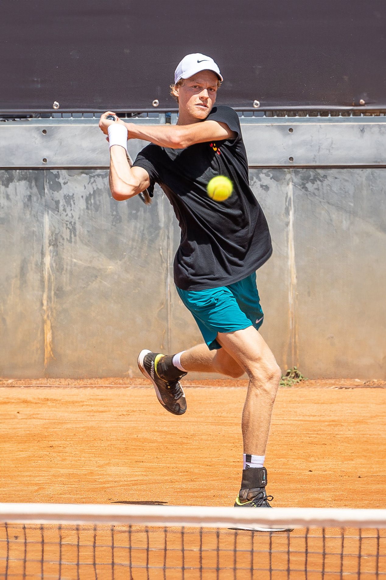 Tennis player in black shirt and teal shorts hits backhand shot on clay court.