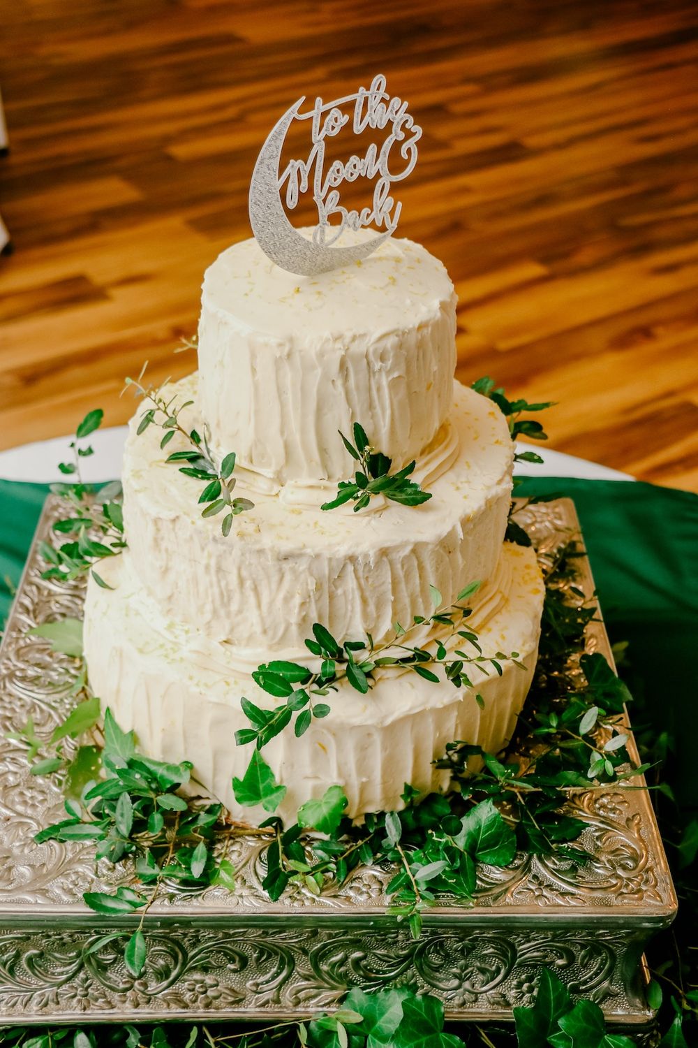 Three-tiered white wedding cake decorated with green foliage and silver moon topper.