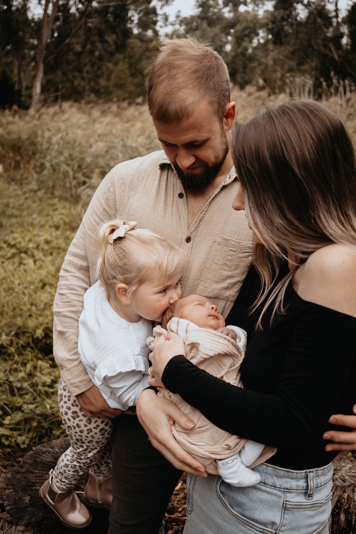 Mum and dad hugging while their daughter kisses her newborn brothers head.