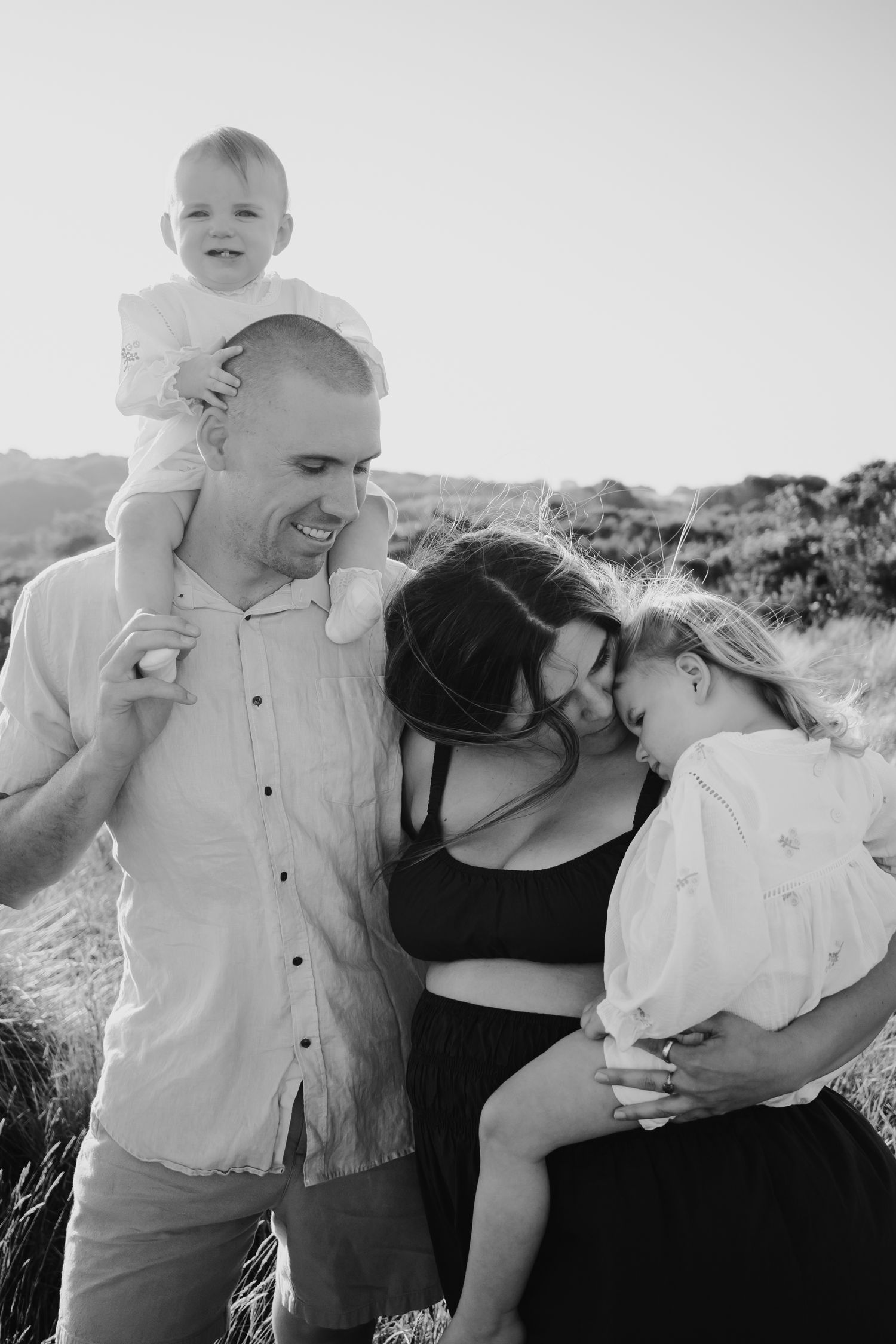 Baby on dad's shoulders while mum carries and kisses toddlers head.