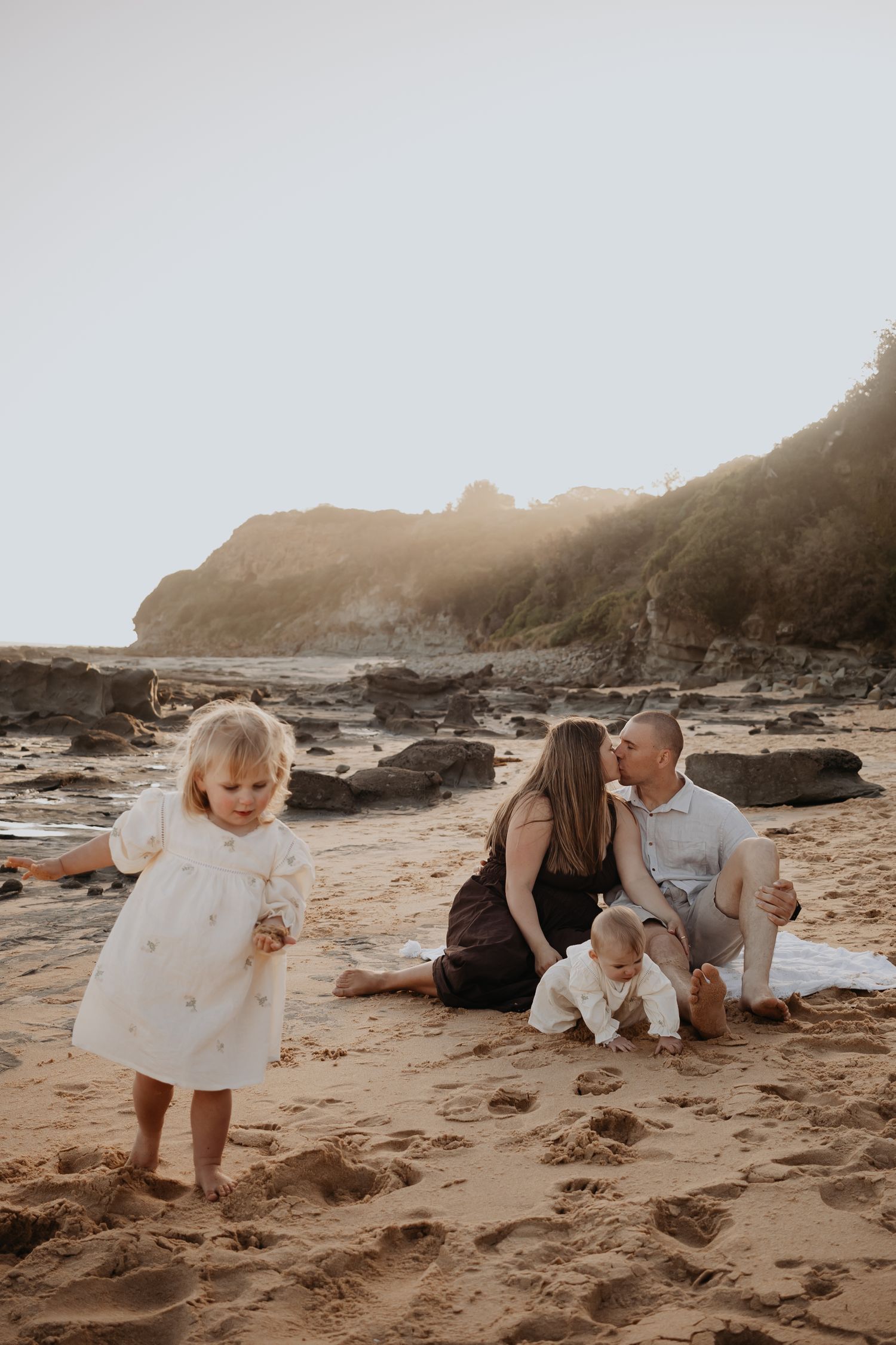 Mum and dad sitting on the beach kissing while other daughter runs off in the sand.