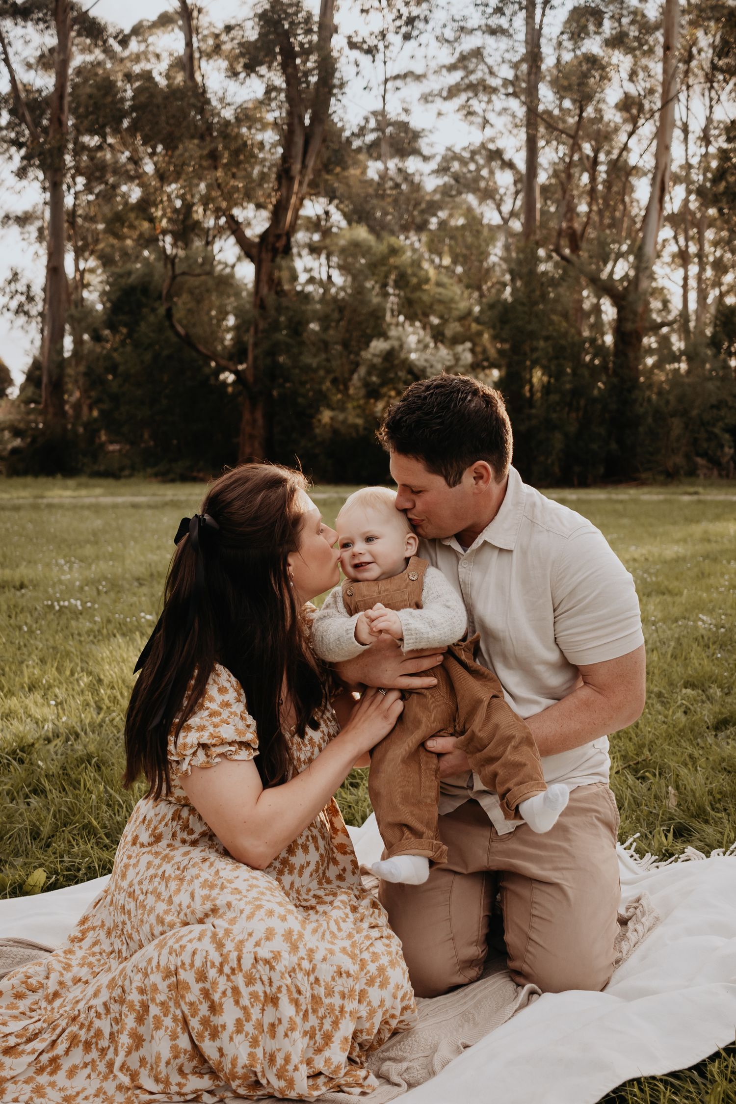 Mum and dad sitting on the grass kissing either side of baby's cheeks.