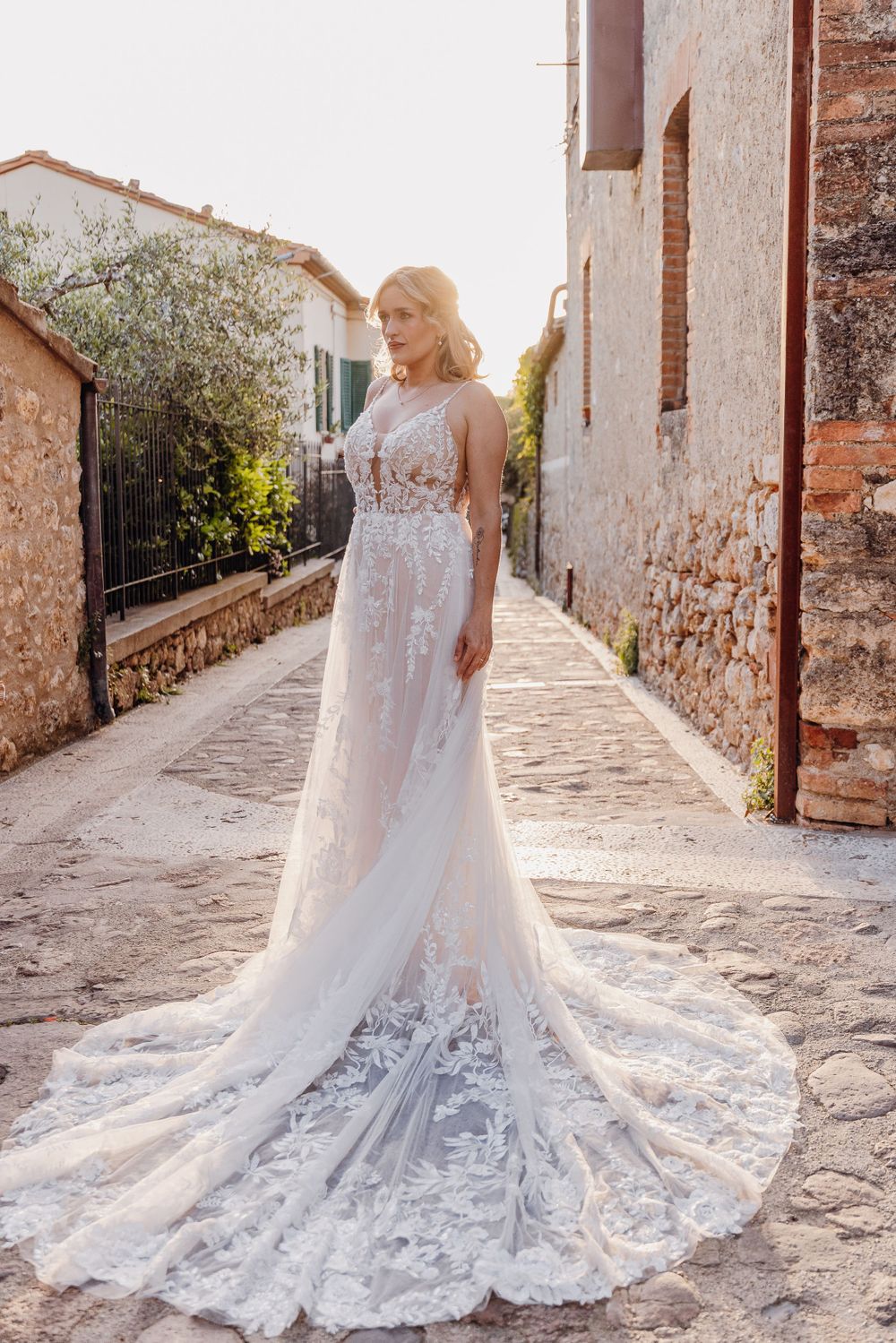 Elegant bride in lace wedding gown with long train poses on rustic Italian street at sunset with stone buildings.