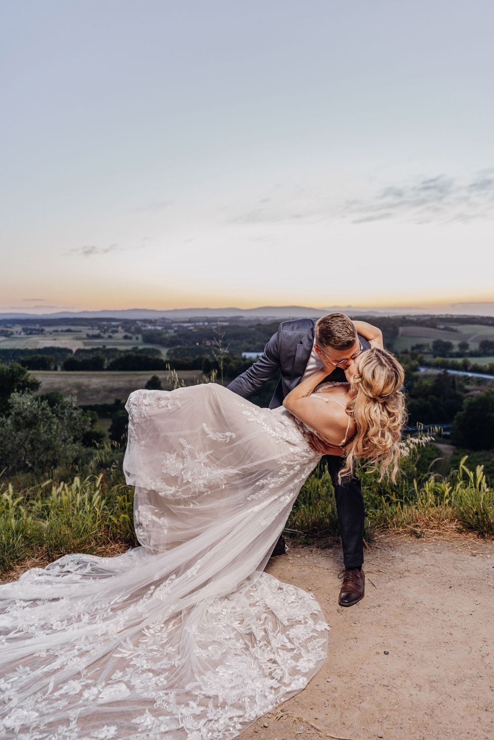 A couple in wedding attire share a romantic dip kiss at sunset overlooking a scenic valley landscape.