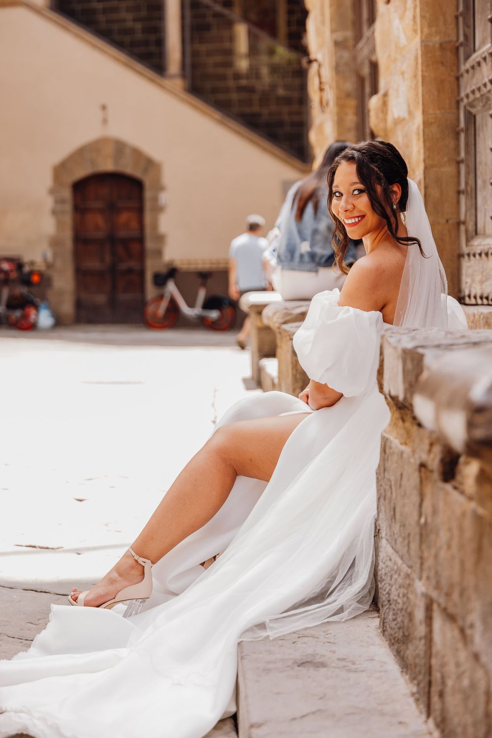 A bride in an off-shoulder white wedding dress poses on stone steps in warm sunlit European architecture.