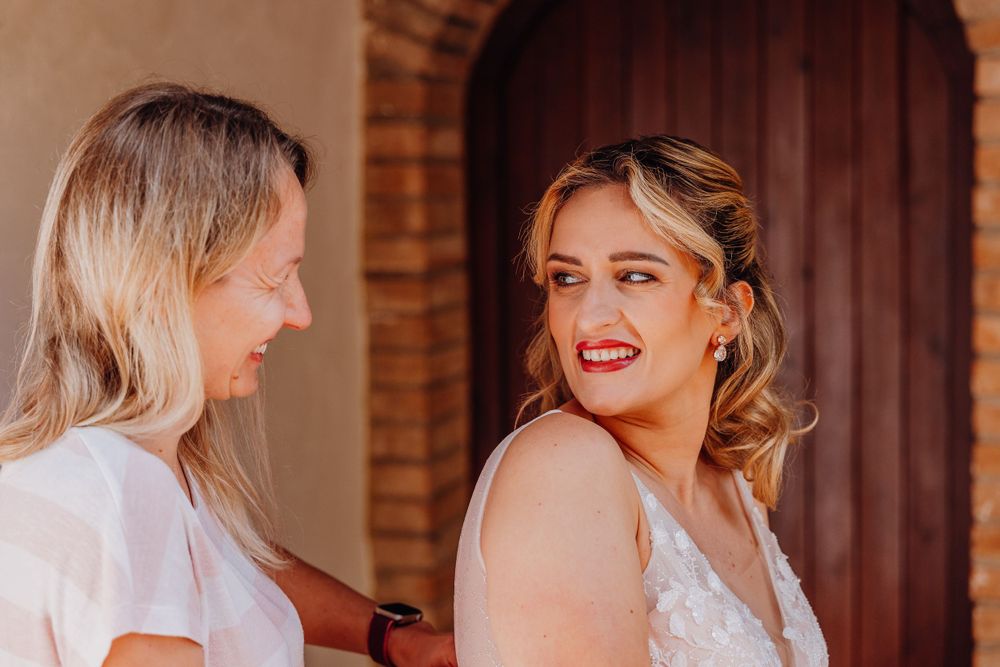 Two women share a joyful moment at a wedding, one wearing white and one wearing a traditional white bridal gown.