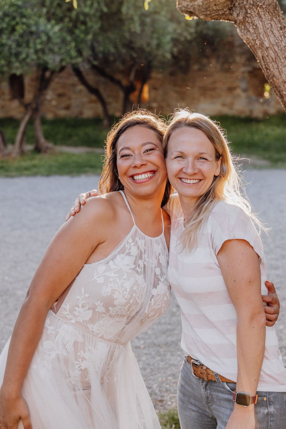 Two friends embrace and smile together outdoors at sunset in a garden setting while wearing white and casual clothing.