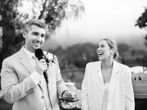 Groomsmen in light colored suits share laughter and drinks during an outdoor wedding celebration.