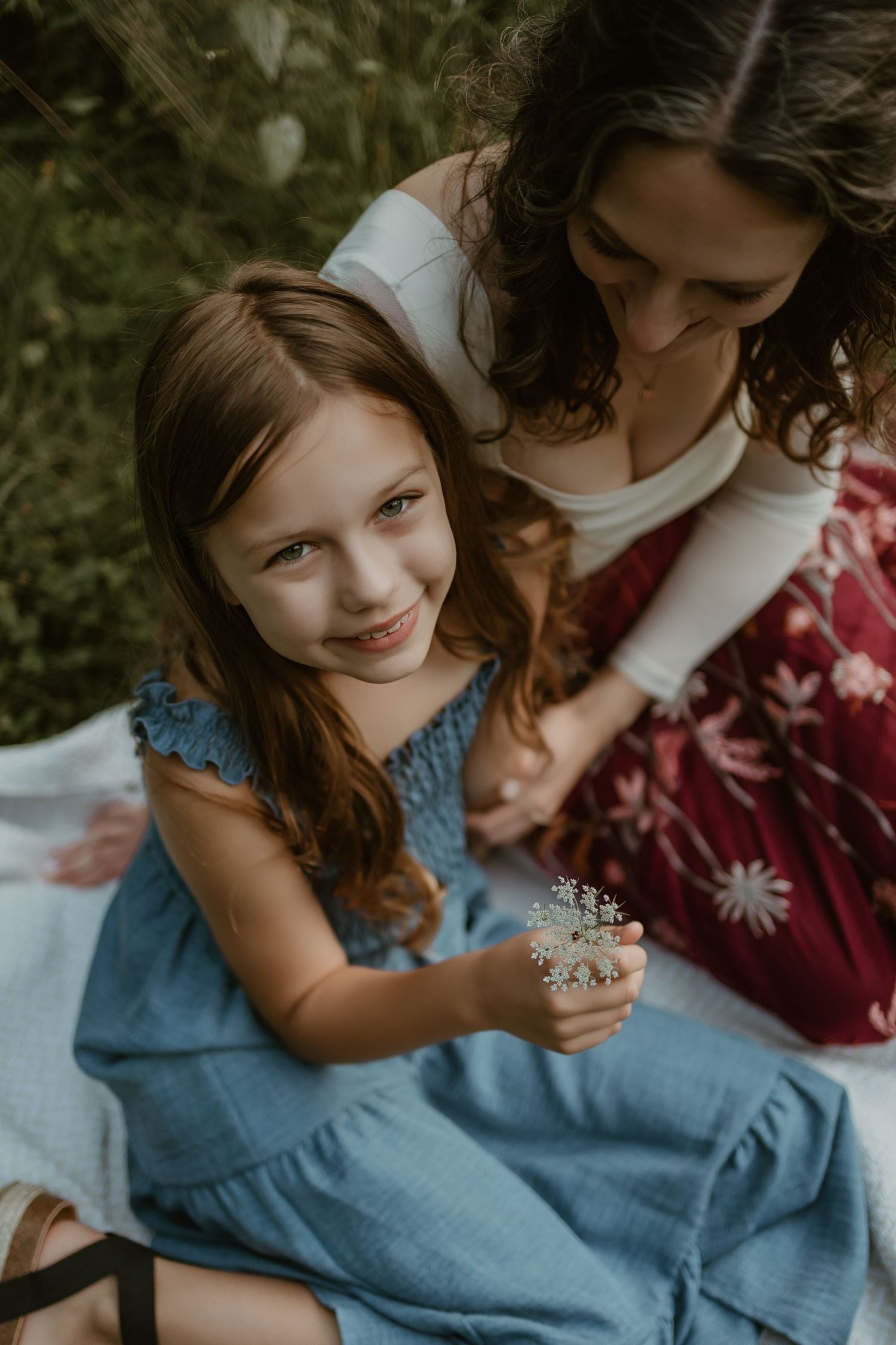 A parent and child share an intimate moment together on a blanket outdoors, wearing blue and burgundy clothing.