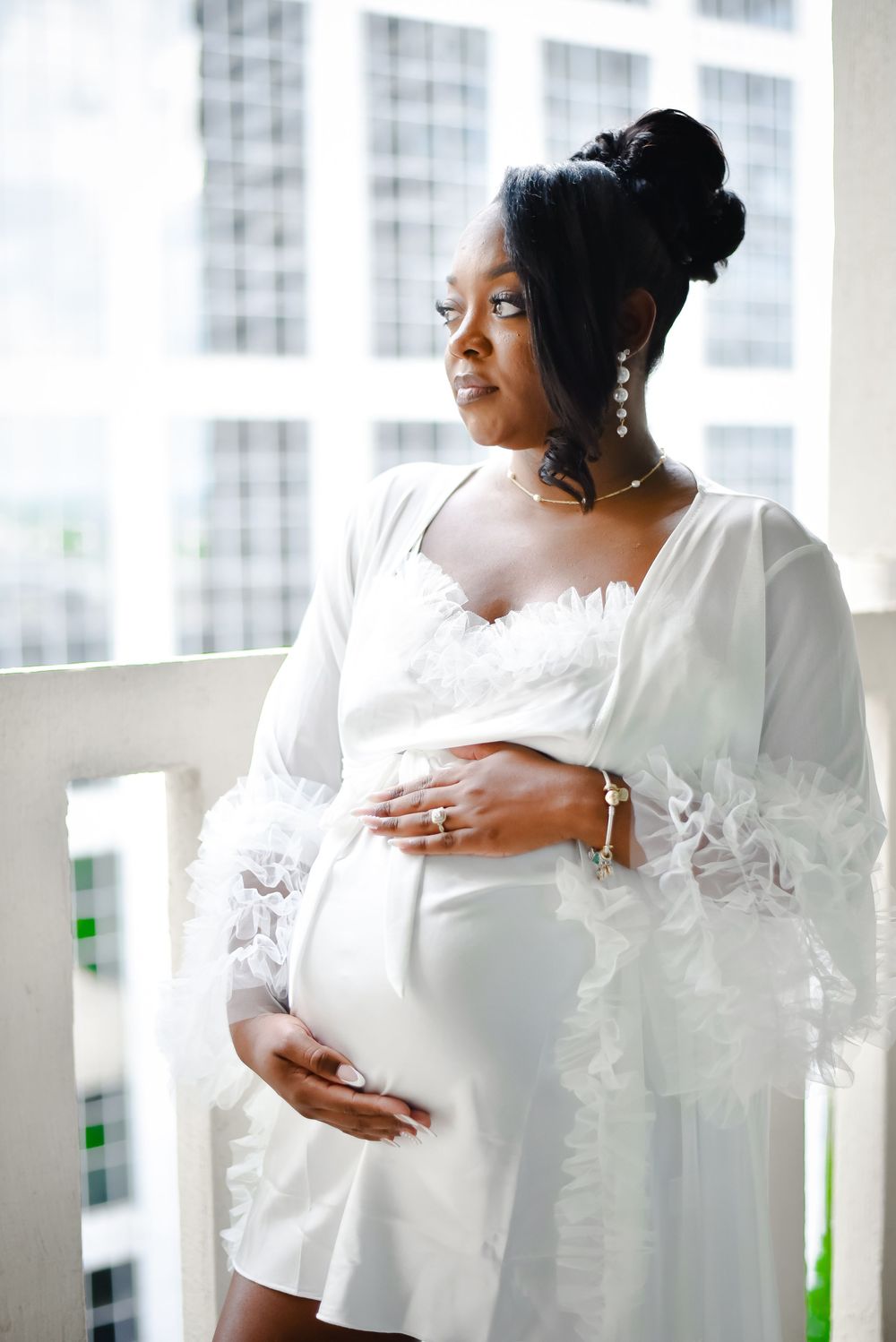 An elegant maternity portrait by a window featuring a white flowing dress.