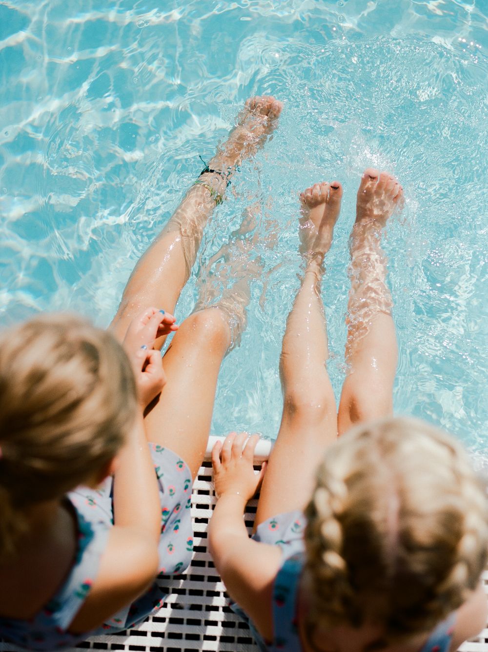 Two young children with braided hair sit at pool edge splashing their legs in clear blue water on a sunny day.