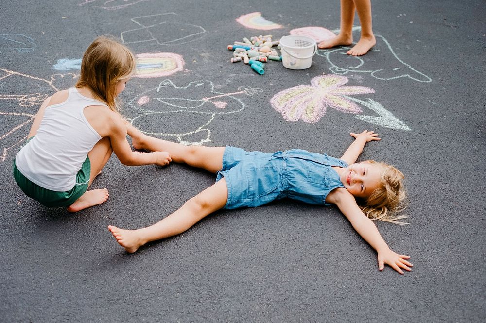 Two children drawing with chalk on asphalt pavement, one lying down beside colorful butterfly and flower artwork.