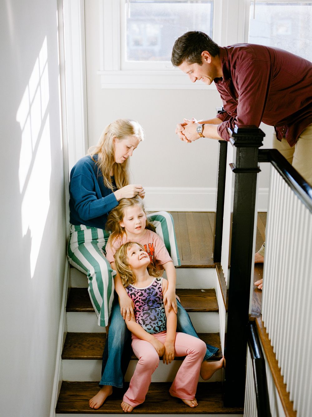 Family on staircase, mother braiding daughter's hair while girls sit on steps and father leans over railing smiling.