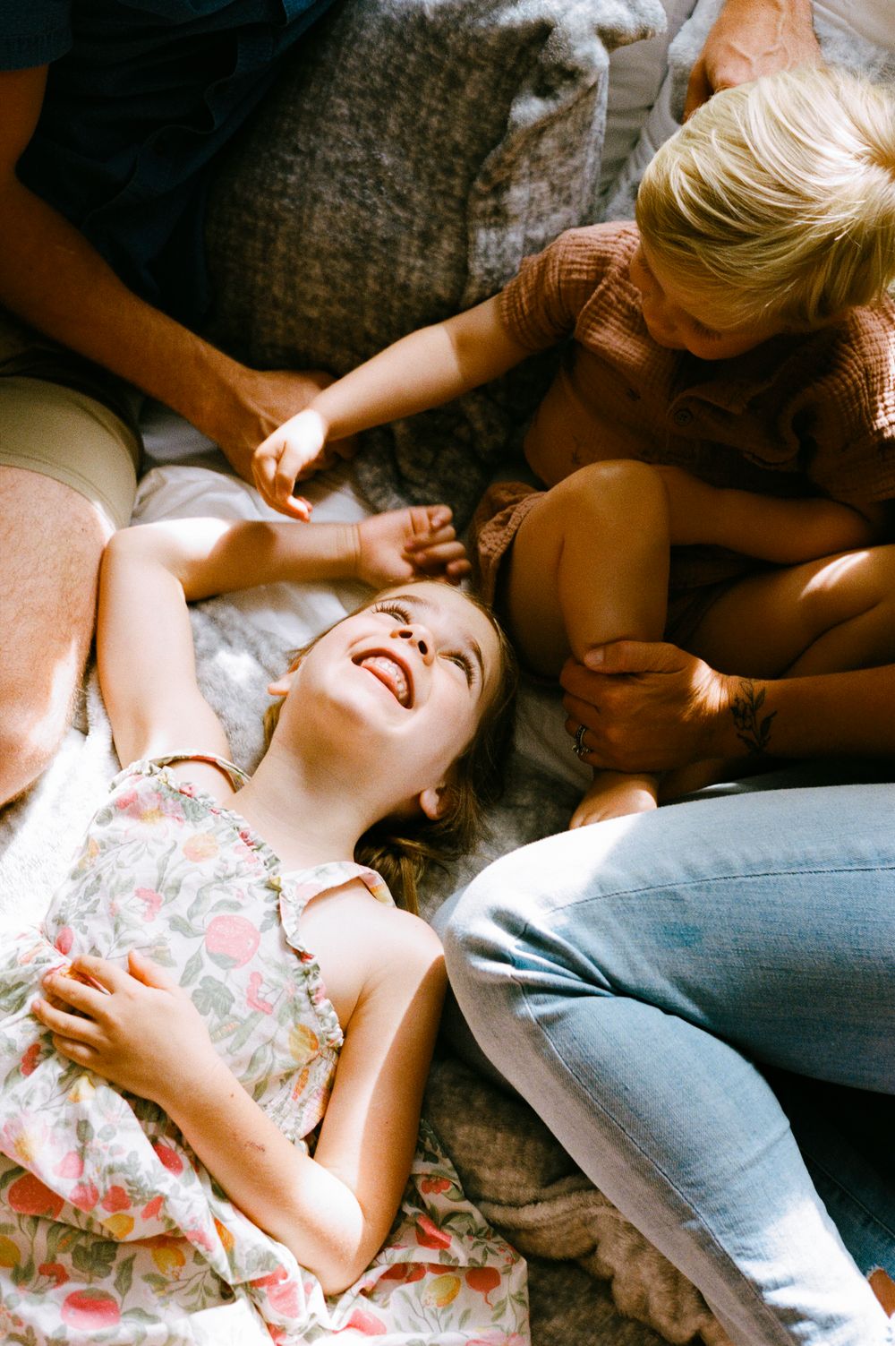 A laughing girl lies on a bed surrounded by family members in a joyful, candid moment of togetherness.