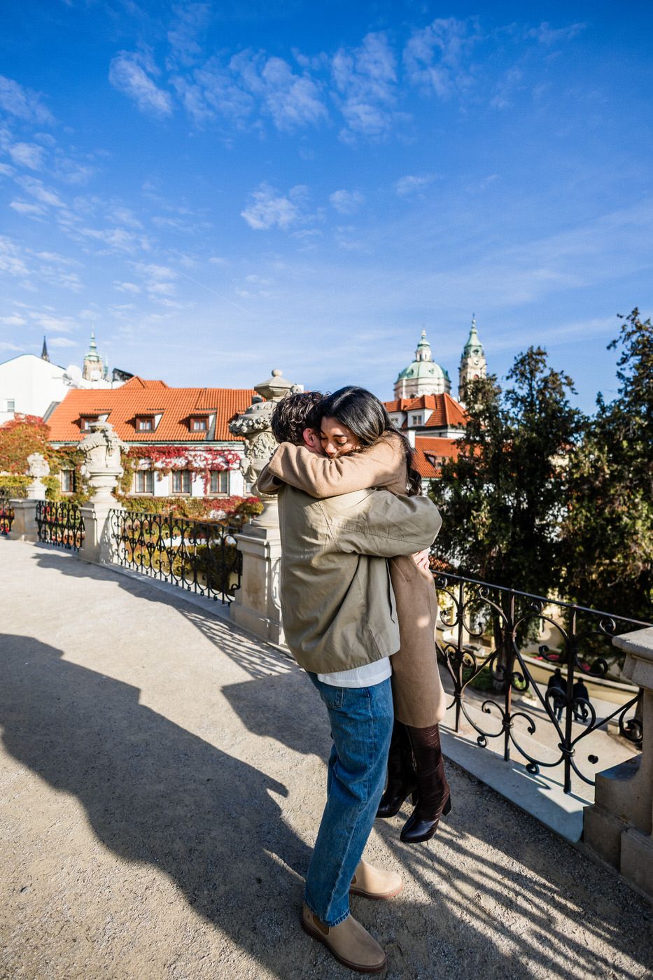 A couple embracing on a scenic terrace with Prague's historic architecture and orange rooftops in the background.