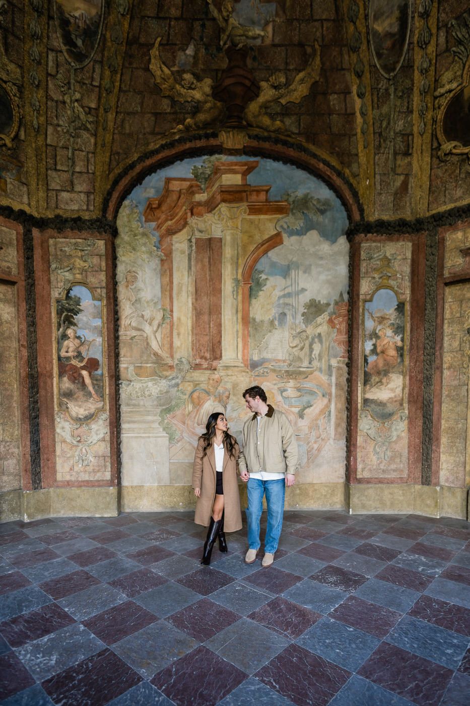 A couple stands beneath an ornate archway with classical frescoes in a historic European building.
