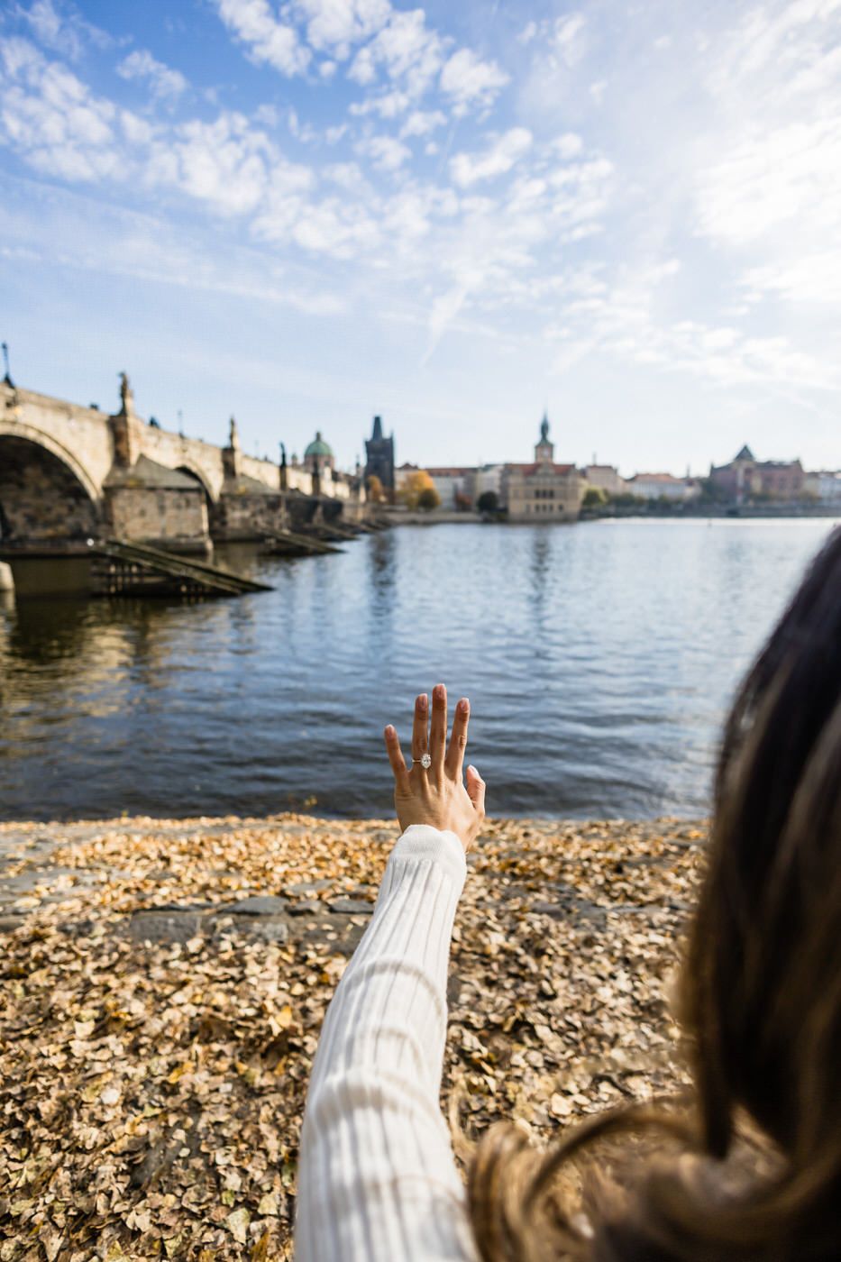 A hand reaches towards the iconic Charles Bridge and Prague skyline along the Vltava River on a sunny day.