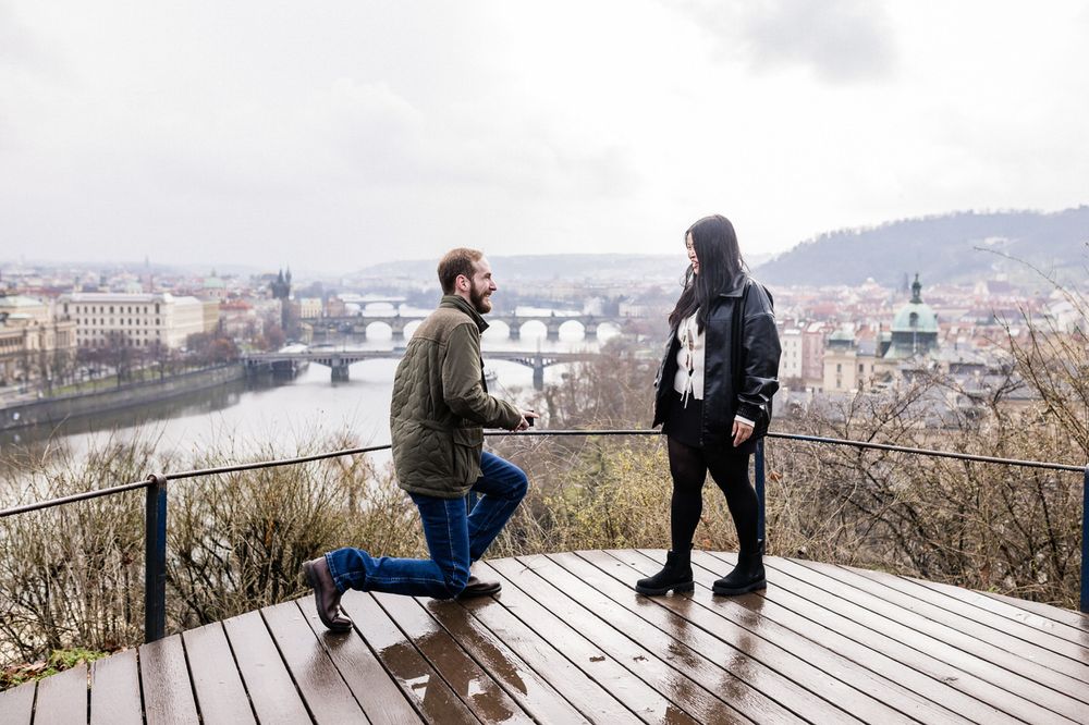 Two people share a special moment on a wooden viewing platform overlooking Prague and its famous bridges on a cloudy day.