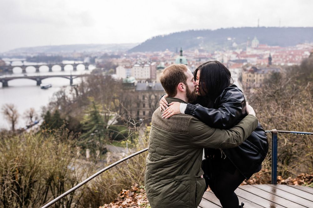 Two people embrace while looking out over a scenic city view from an elevated viewpoint on a cloudy day.