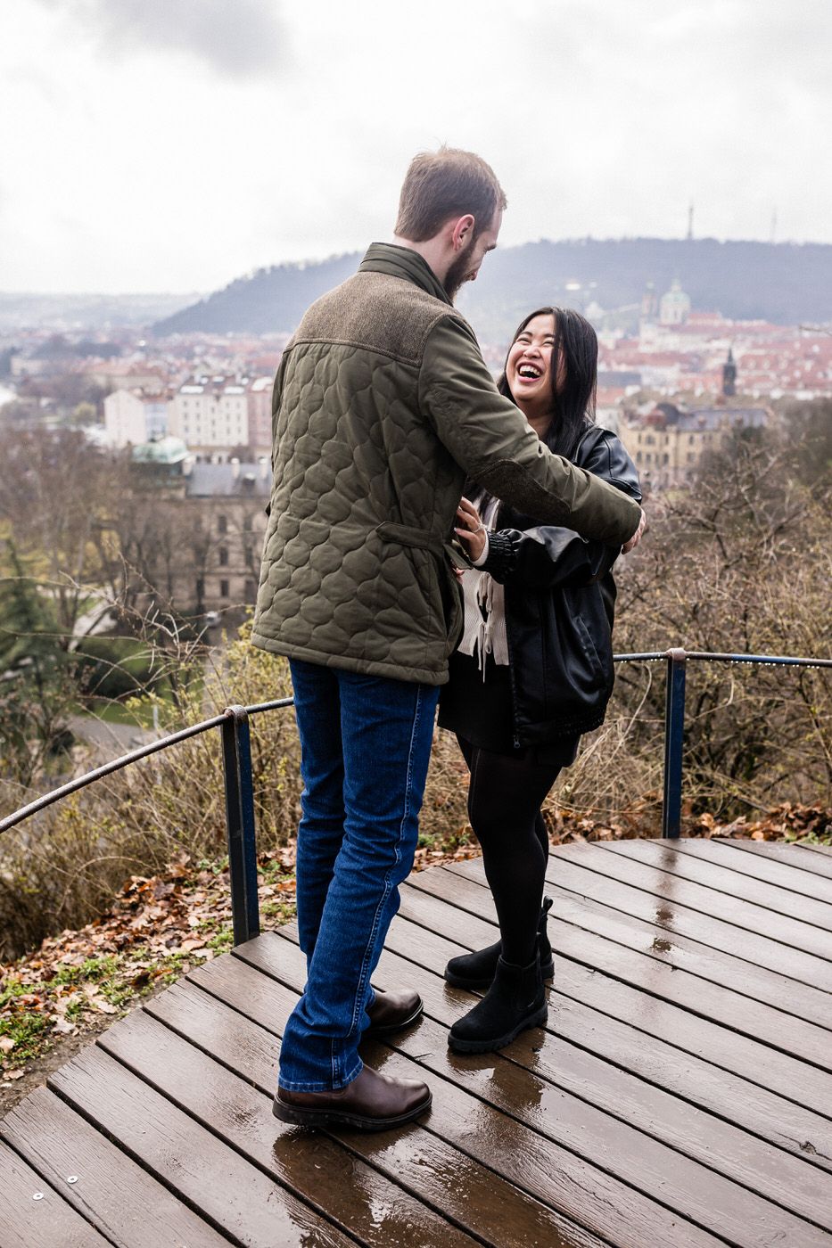 A couple embracing on a wooden observation deck overlooking a European cityscape on a cloudy day.