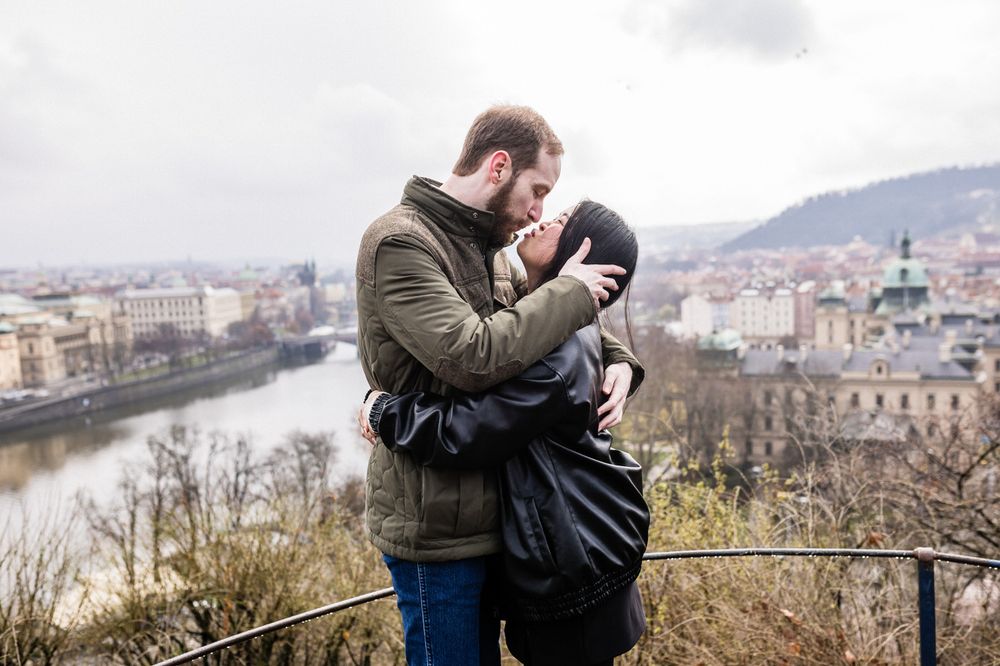 Two people embrace while standing on an overlook with a scenic river and cityscape view in the background.