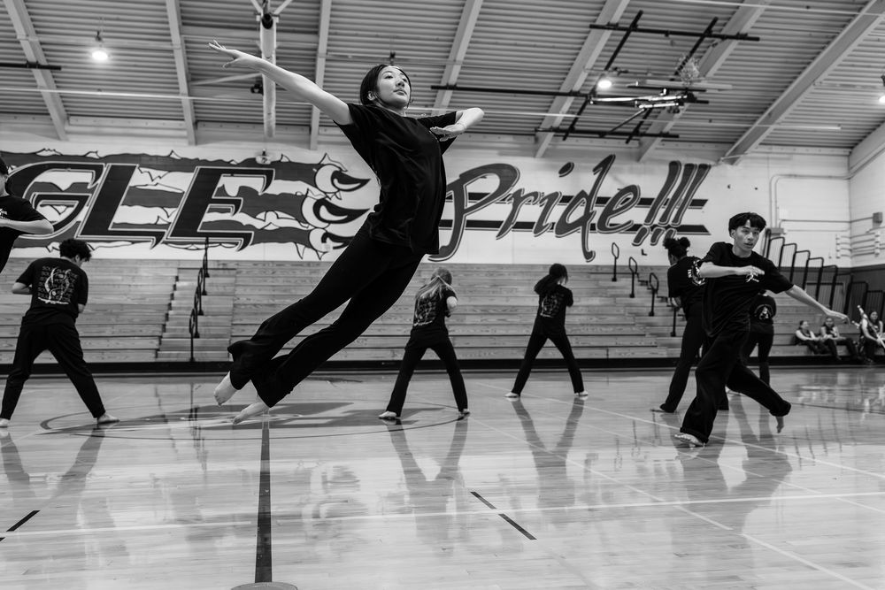Dancers leap and move gracefully across a gymnasium floor during dance practice in black and white photography.