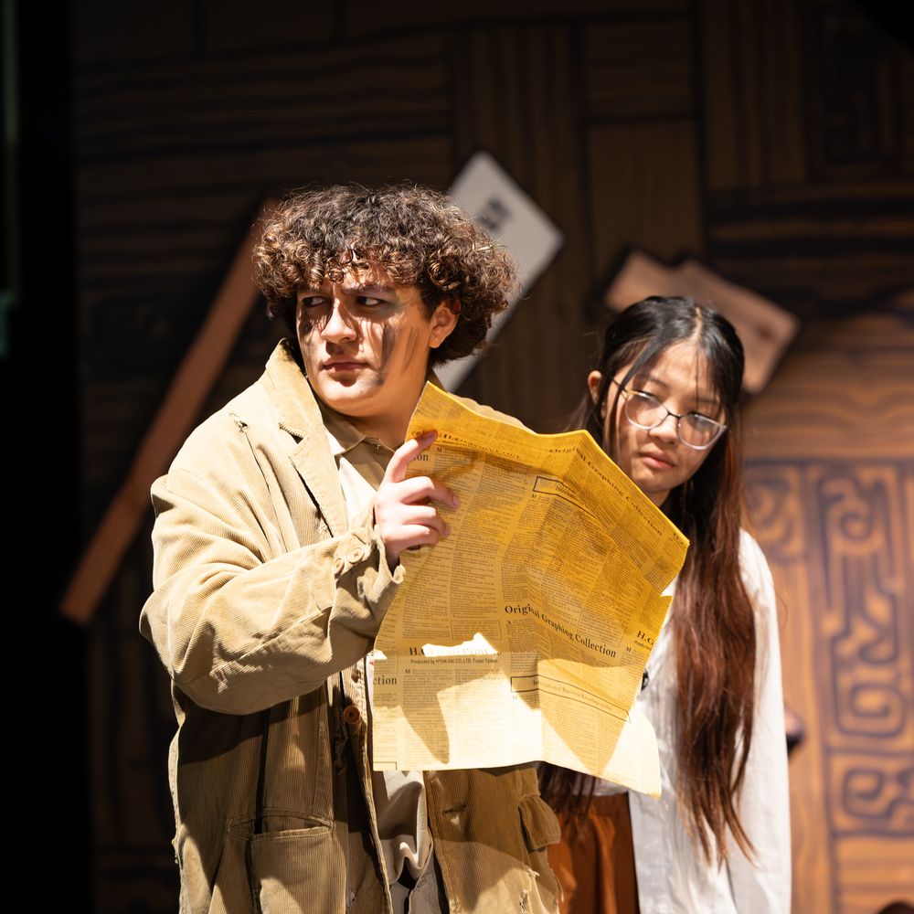 Two young theater actors on stage, one holding a large aged map or document during a dramatic performance.