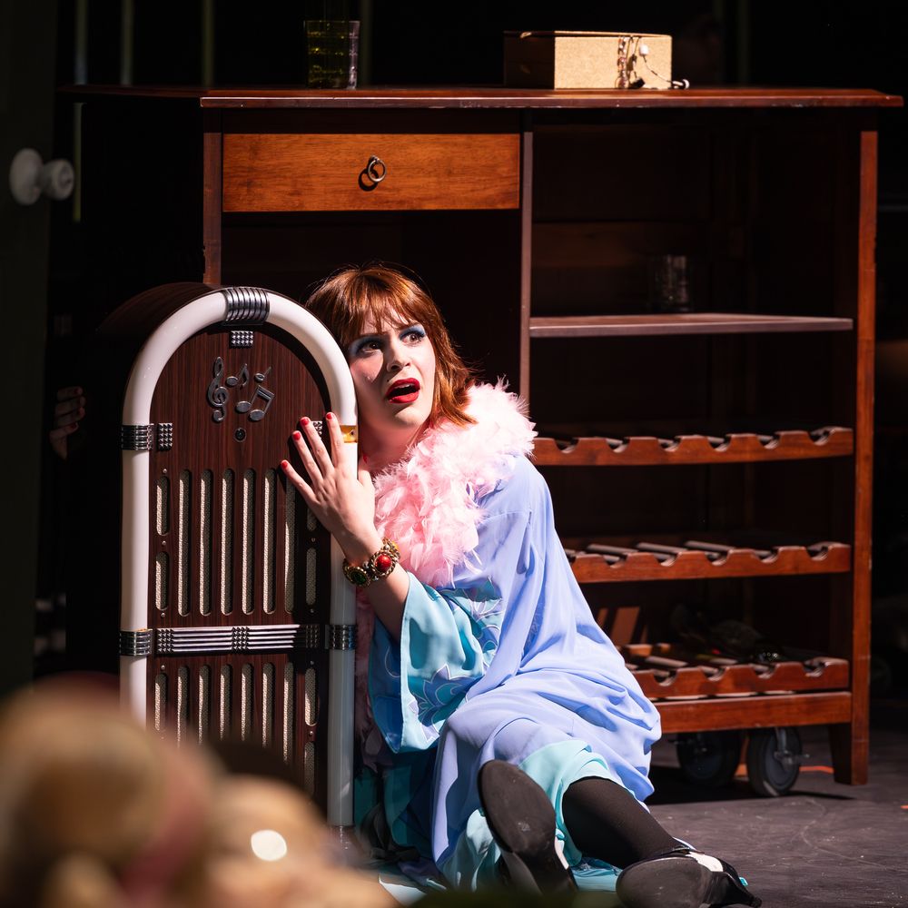 Actress in blue robe with pink feather boa performs dramatically beside vintage jukebox on theater stage.