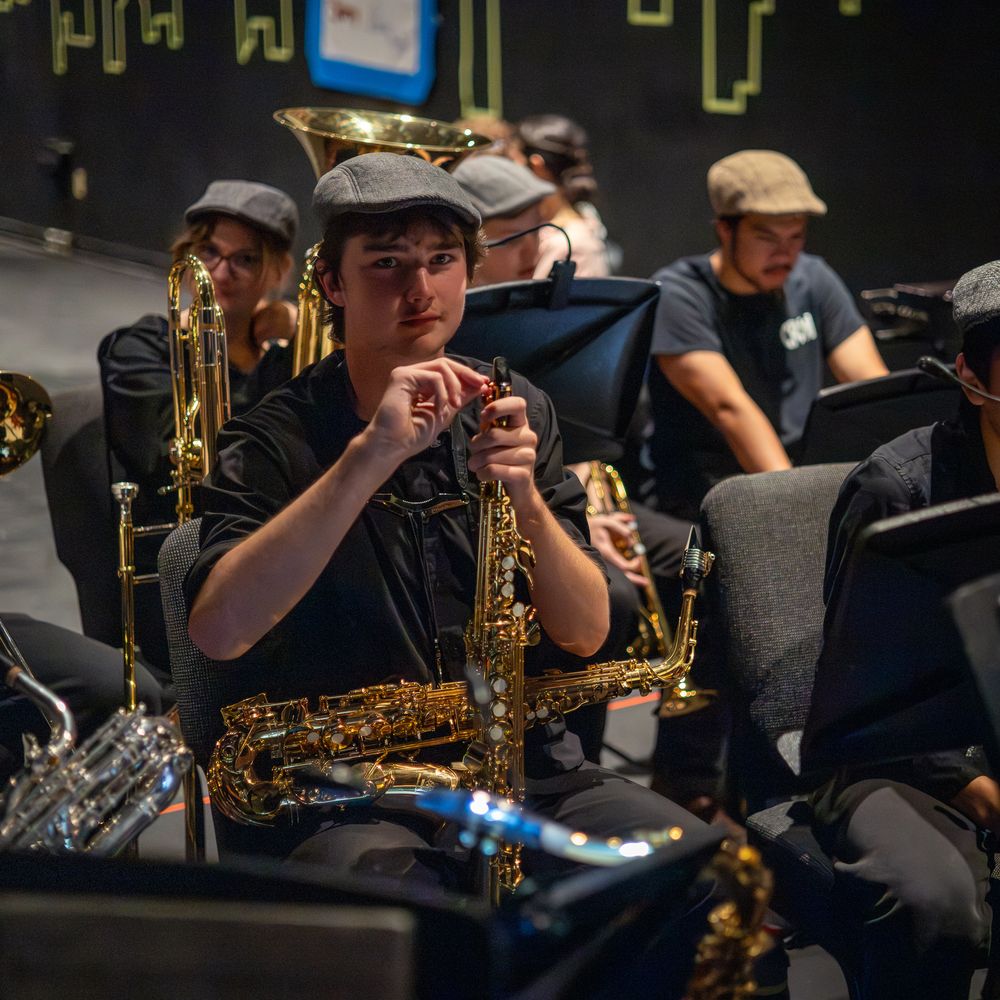 Young musician in a newsboy cap holds a saxophone during a jazz band rehearsal with brass instruments.