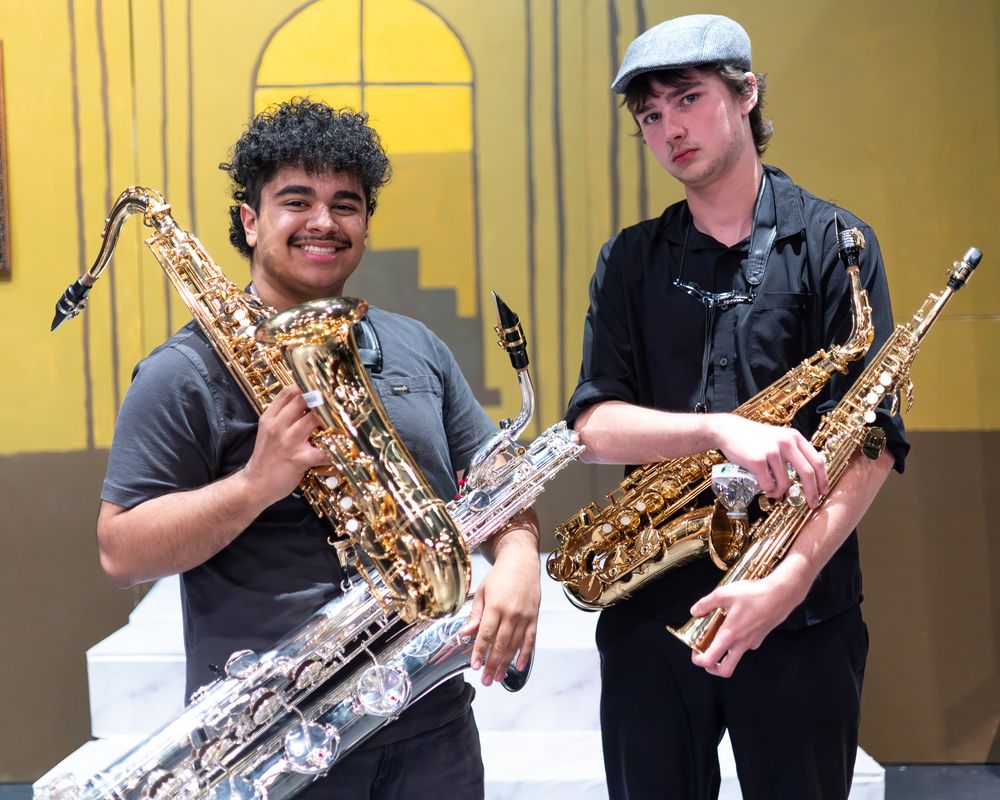 Two young musicians smiling and holding saxophones in front of a yellow artistic backdrop.