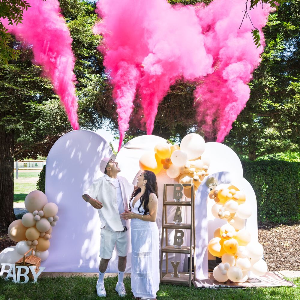 Couple celebrates gender reveal with pink smoke cannons at decorated outdoor baby shower with balloon arch.