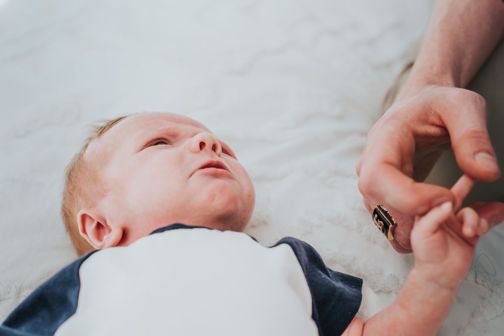 A newborn lays looking up and holding an adults hand with their tiny fingers.