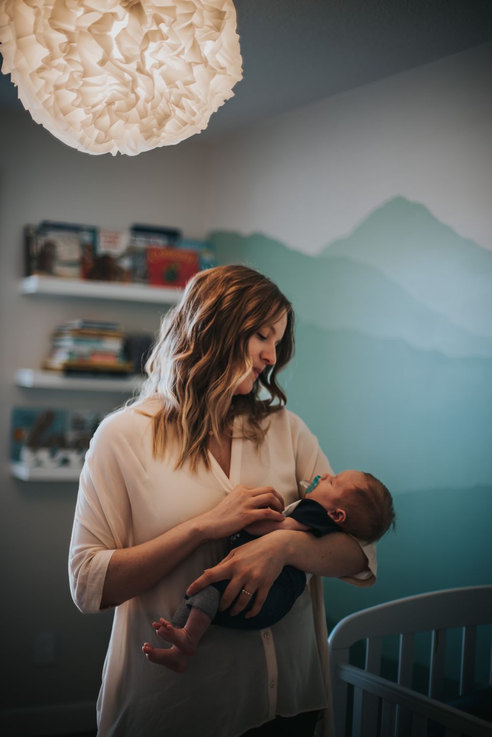 Woman stands holding her newborn in a beautifully decorated nursery with a mountain mural on the wall.
