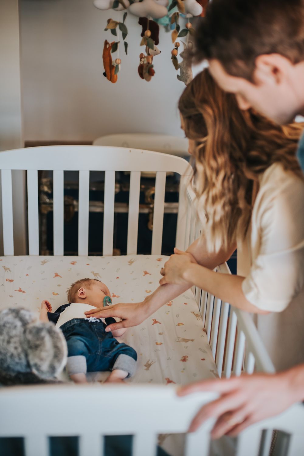 Mom and dad lean over their white crib to hold their baby's hand.
