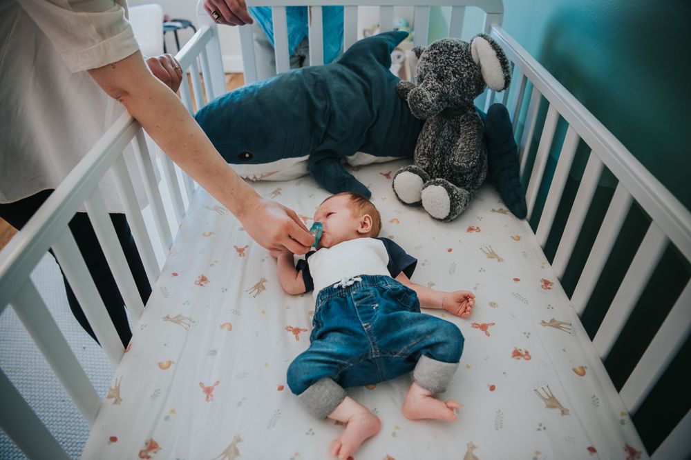A mother's hand reaches down to hold her baby's hand as he lays in his crib with stuffed animals.