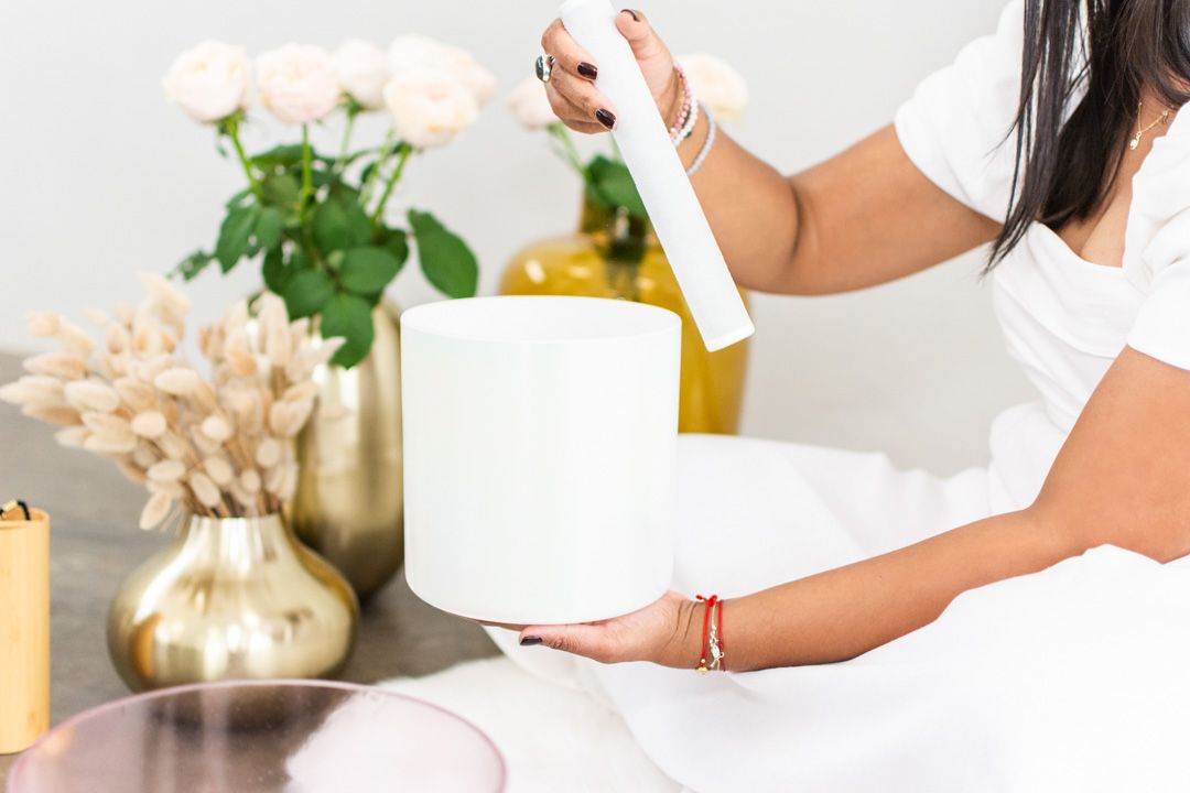 Hands holding white sound bowl with roses and dried flowers in gold vase on counter.