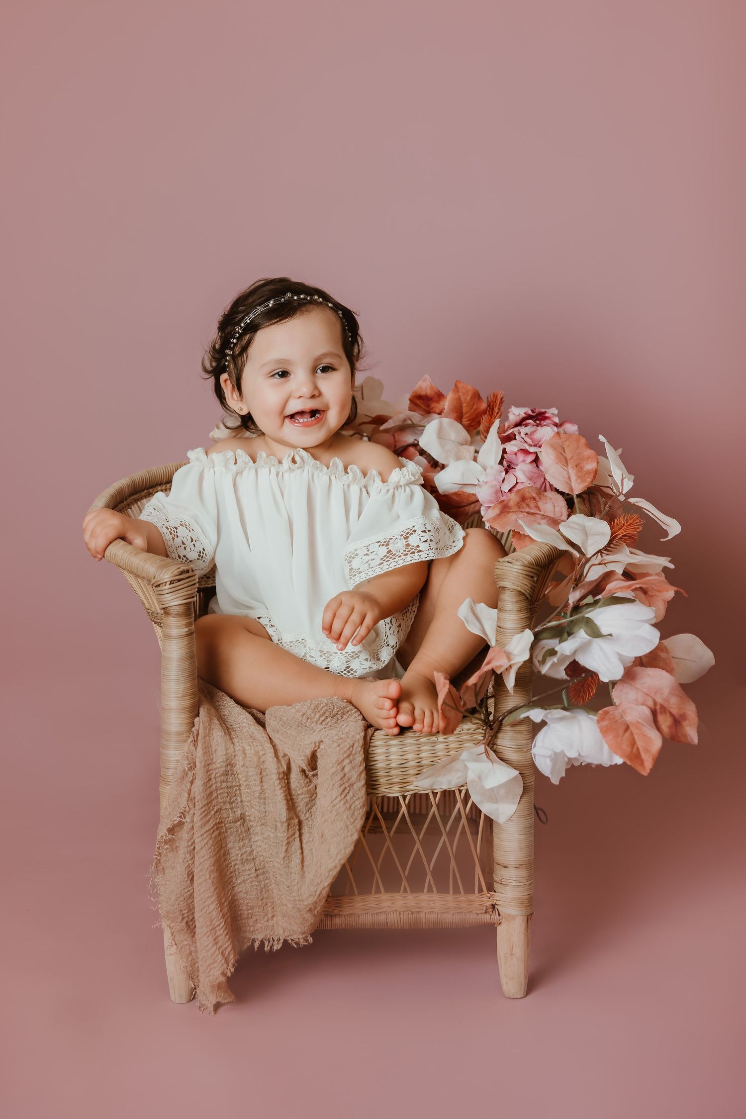 Portrait of young child in white lace dress sitting on wicker chair with pink floral decorations.