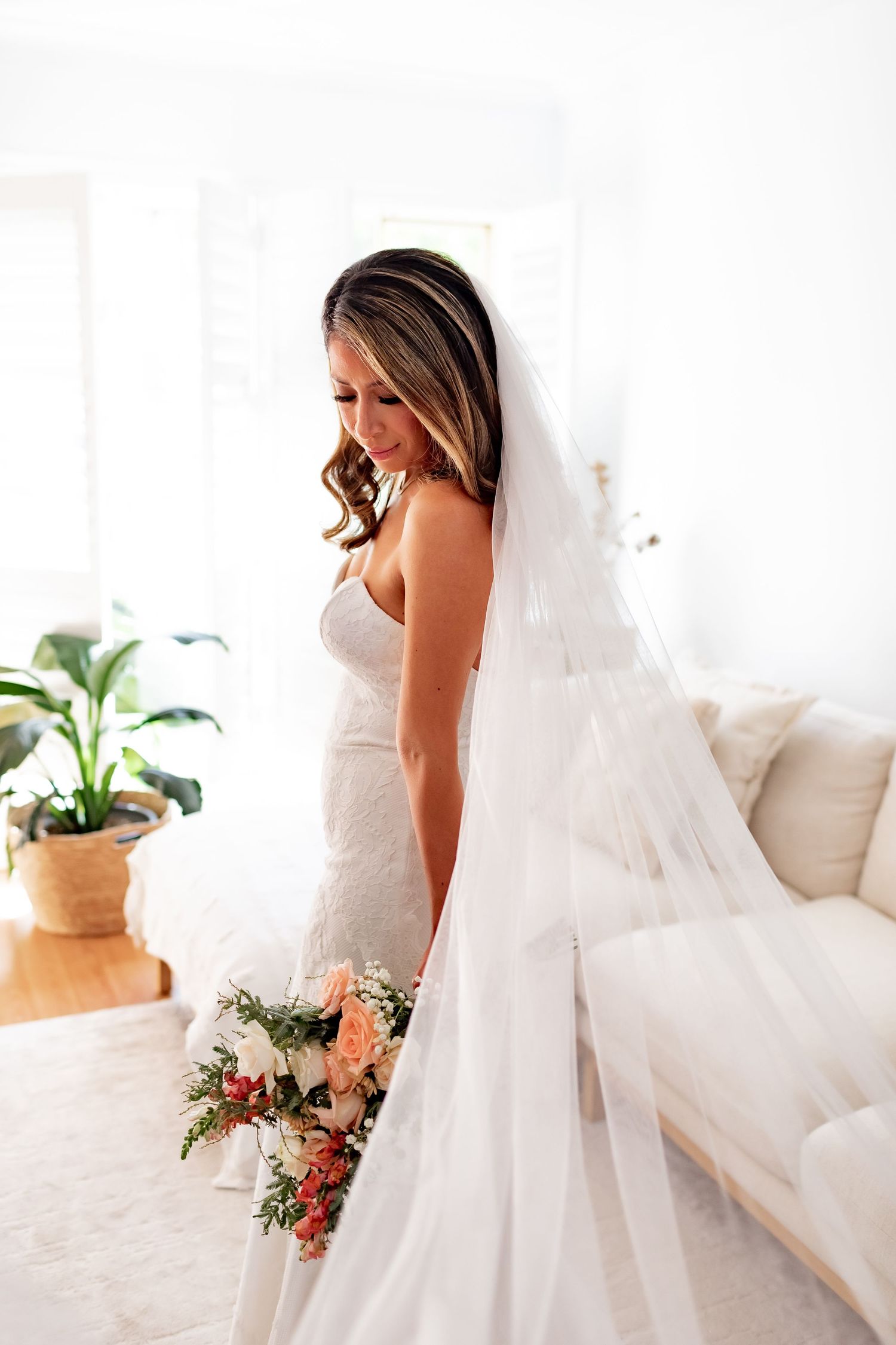Elegant bride in white dress with flowing veil holds pink and white bouquet in bright room.