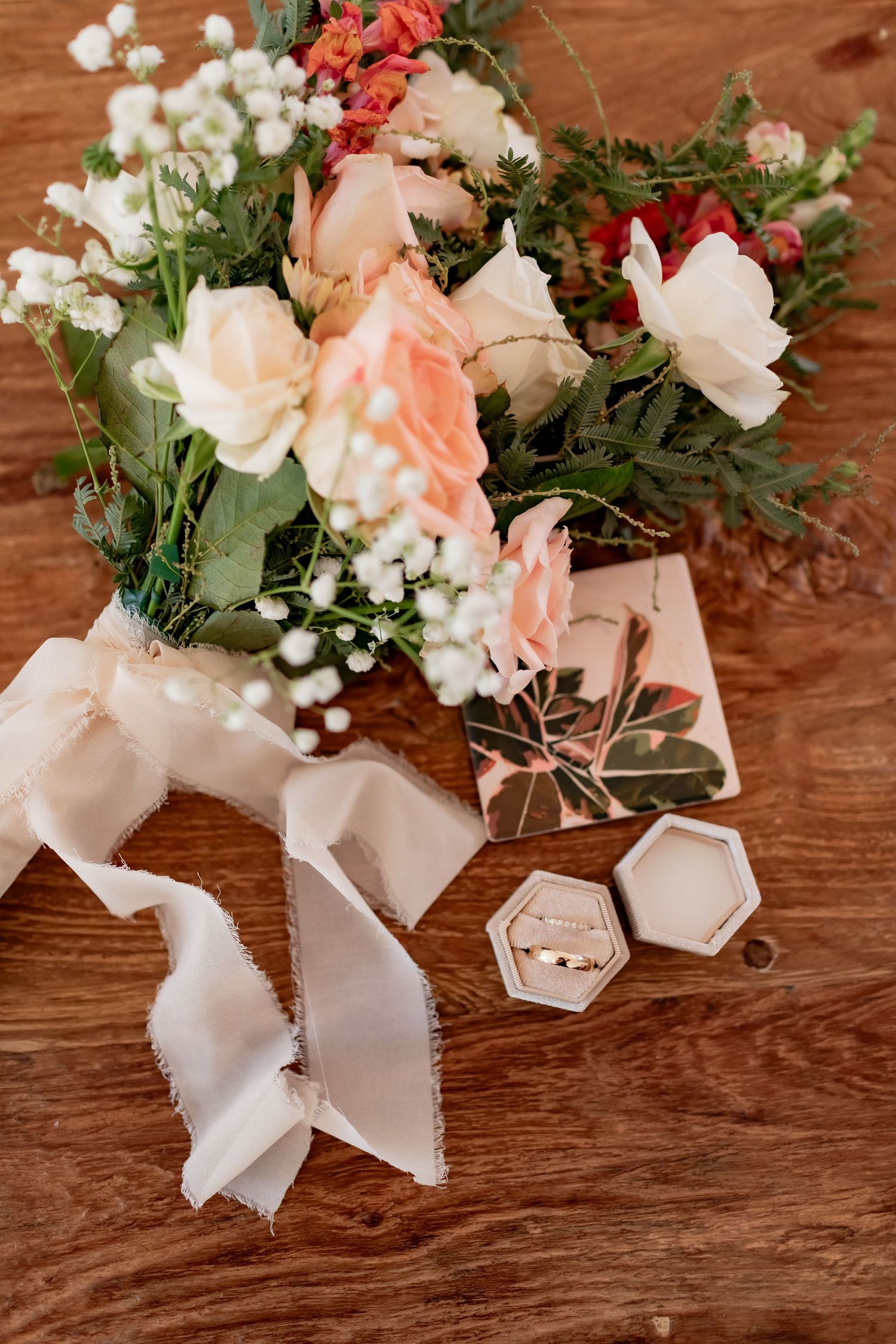 Wedding bouquet with coral and white flowers alongside hexagonal ring boxes on wooden surface.