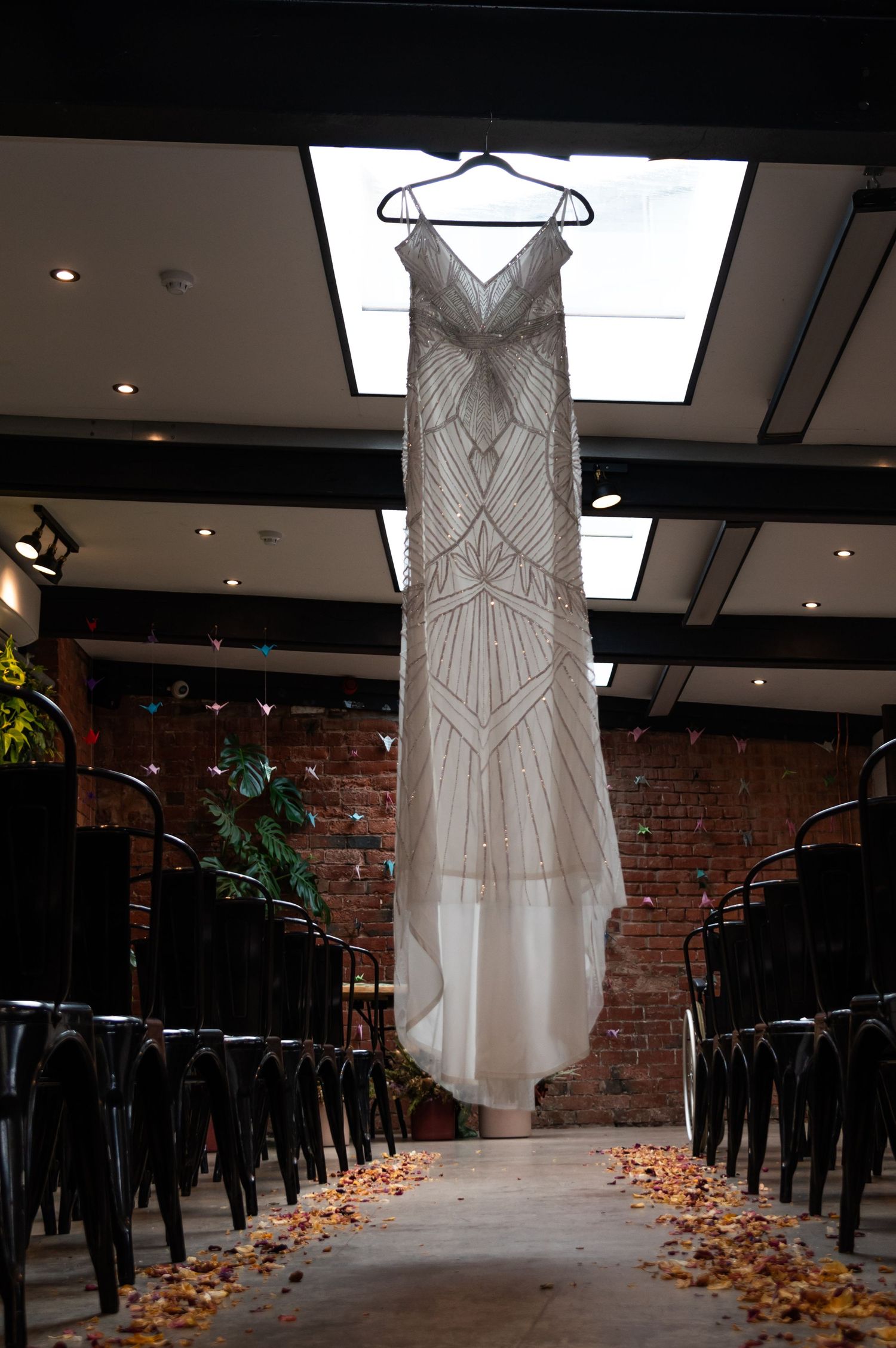 White beaded wedding dress hanging in a rustic brick venue with scattered rose petals on black chairs.