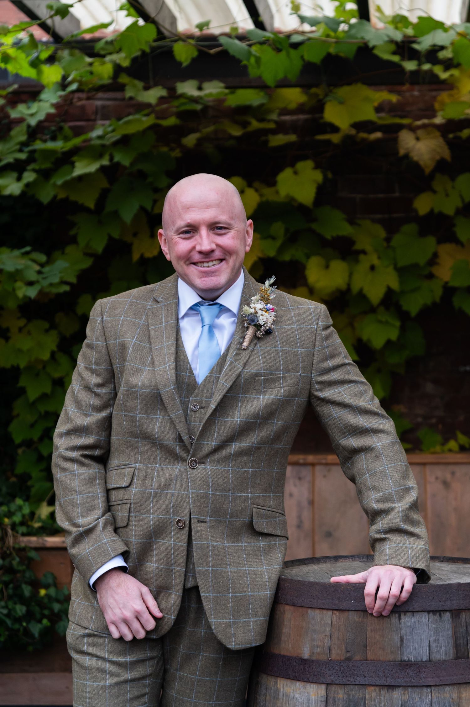 Groom in checkered tweed suit posing against green foliage backdrop.