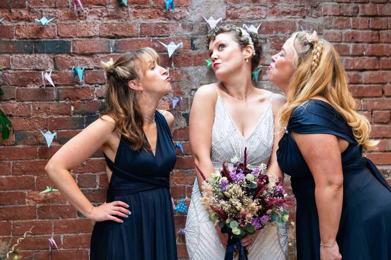 Bridesmaids in navy dresses posing against brick wall with bridal bouquet.
