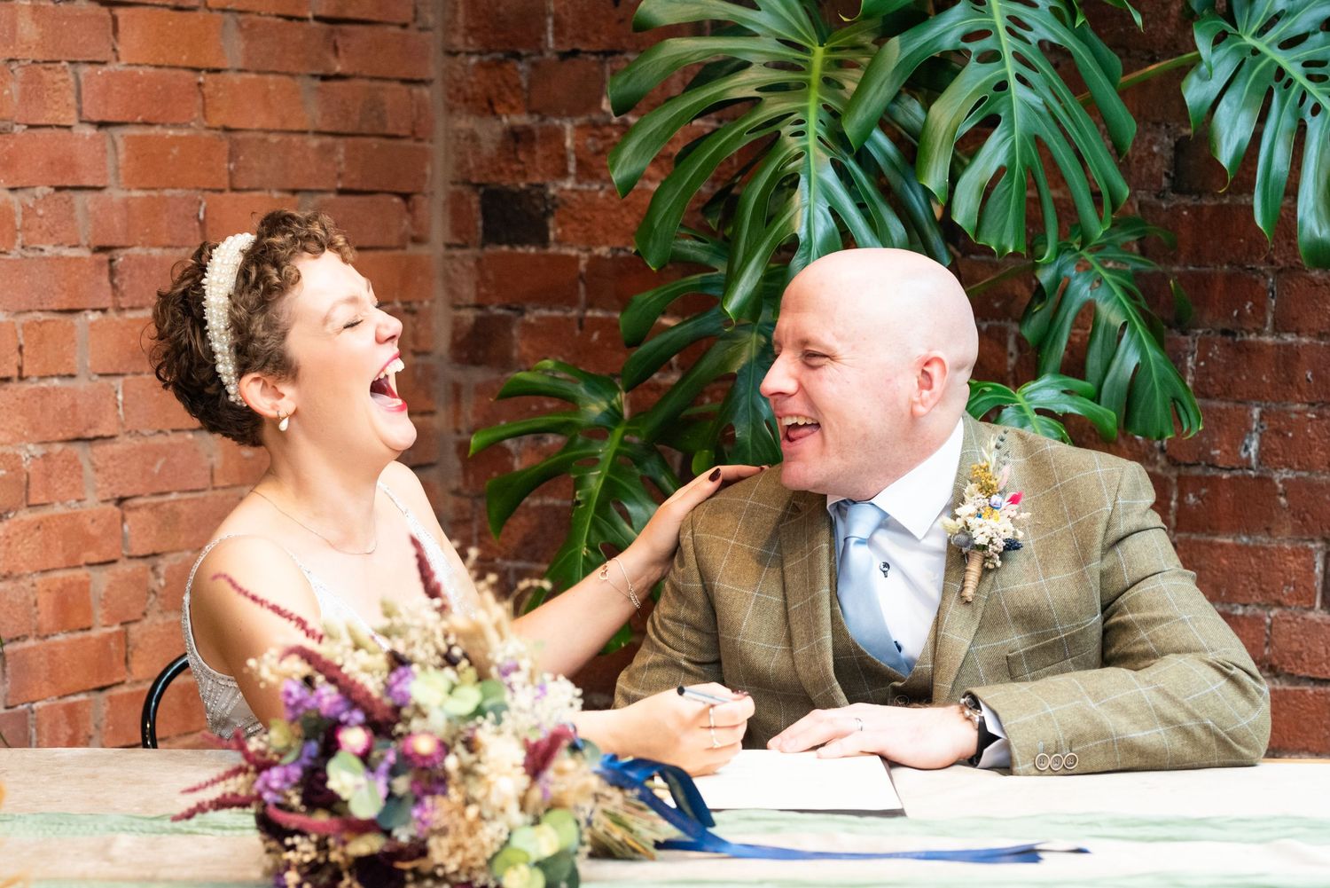 Couple sharing a candid moment of laughter against brick wall with tropical plants.
