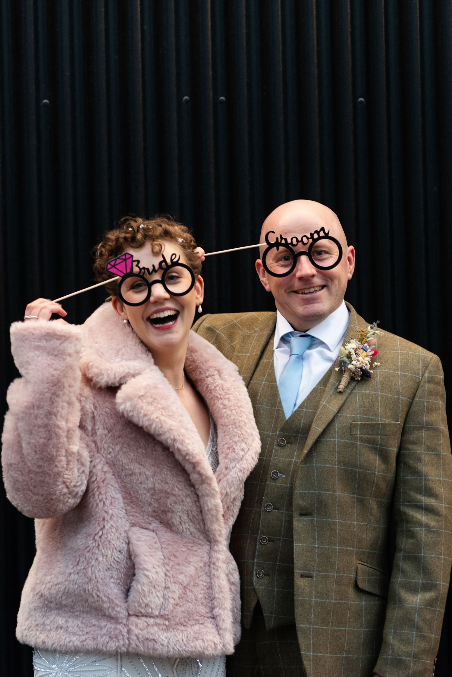 Wedding couple in formal attire with playful glasses poses against dark backdrop.